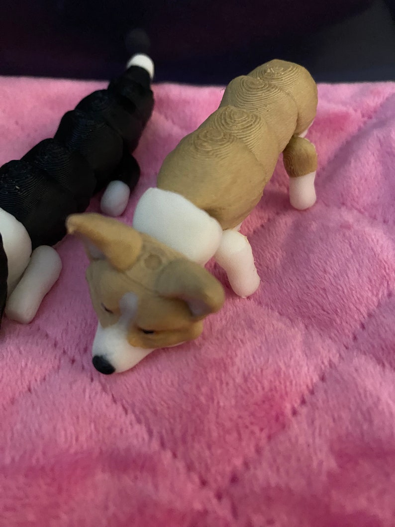 May include: Two 3D printed dog figurines. One is black and white and the other is brown and white. The brown and white dog is lying down with its head resting on the pink surface.