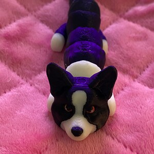May include: A 3D printed black and white corgi dog figurine with a segmented body. The figurine is lying on a pink fuzzy surface.