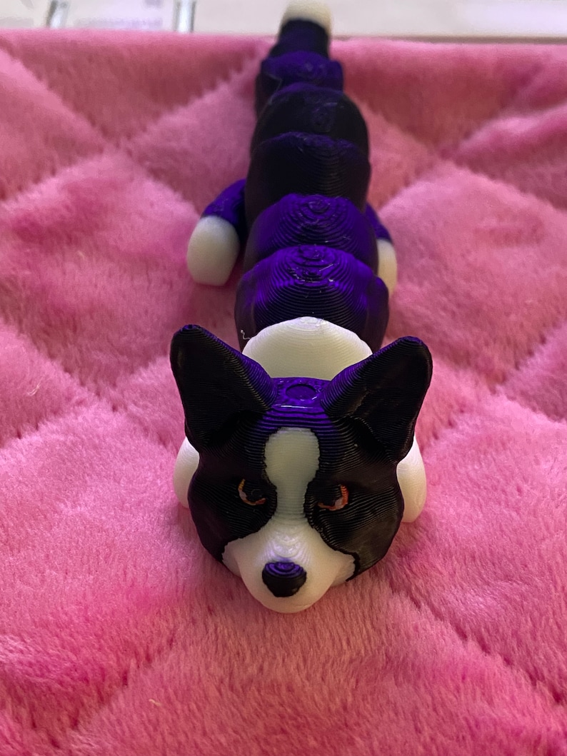 May include: A 3D printed black and white corgi dog figurine with a segmented body. The figurine is lying on a pink fuzzy surface.