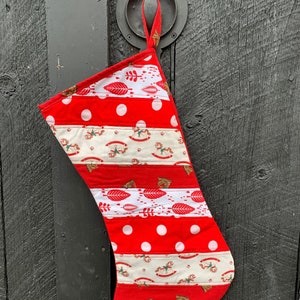 Puede incluir: Una media de Navidad roja y blanca con un diseño de patchwork. La media está hecha de diferentes telas con varios patrones, incluyendo lunares, rayas y hojas. Tiene un lazo en la parte superior para colgarla.
