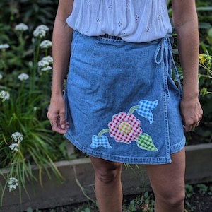 May include: A woman wearing a white eyelet top with a tie at the neck and a blue denim wrap skirt with a colorful floral applique.
