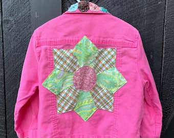 May include: A pink denim jacket with a patchwork flower design on the back. The flower is made of different fabrics, including plaid, paisley, and solid colors. The jacket has a decorative collar with fabric flowers.