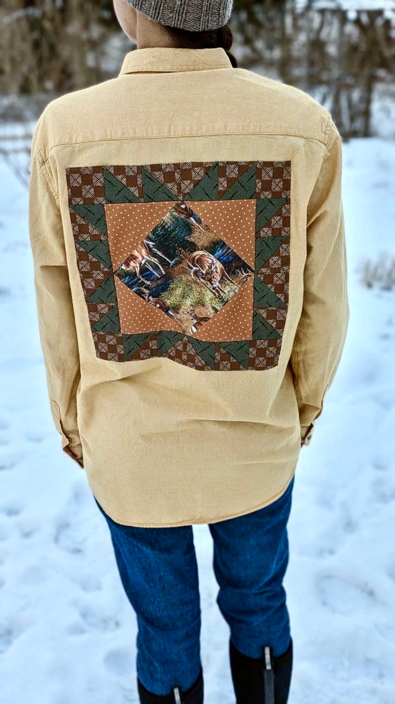Puede incluir: Una camisa de botones de color tostado con un dise&ntilde;o de patchwork en la espalda. El patchwork es un cuadrado con un patr&oacute;n marr&oacute;n, verde y blanco. El centro del patchwork presenta una impresi&oacute;n de ciervos en un entorno de bosque.