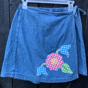 May include: A blue denim wrap skirt with a floral applique. The applique features a red and white gingham flower with a yellow center, blue and white gingham leaves, and green leaves.