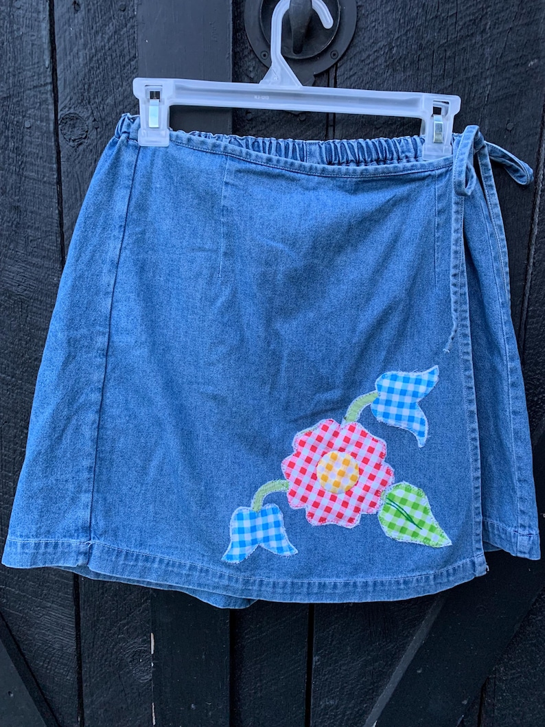 May include: A blue denim wrap skirt with a floral applique. The applique features a red and white gingham flower with a yellow center, blue and white gingham leaves, and green leaves.