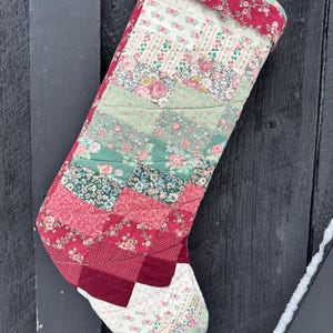 Puede incluir: Un calcetín de Navidad hecho a mano con un diseño de patchwork. El calcetín presenta varios patrones florales en tonos de rojo, rosa, verde y blanco. La parte superior y el puño están adornados con tela floral roja. Colgando de una puerta de madera oscura.