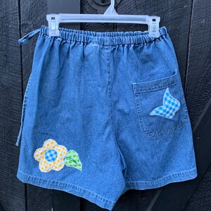 May include: Blue denim shorts with a yellow flower and green leaf applique on the left leg and a blue and white checkered fabric applique on the right back pocket.