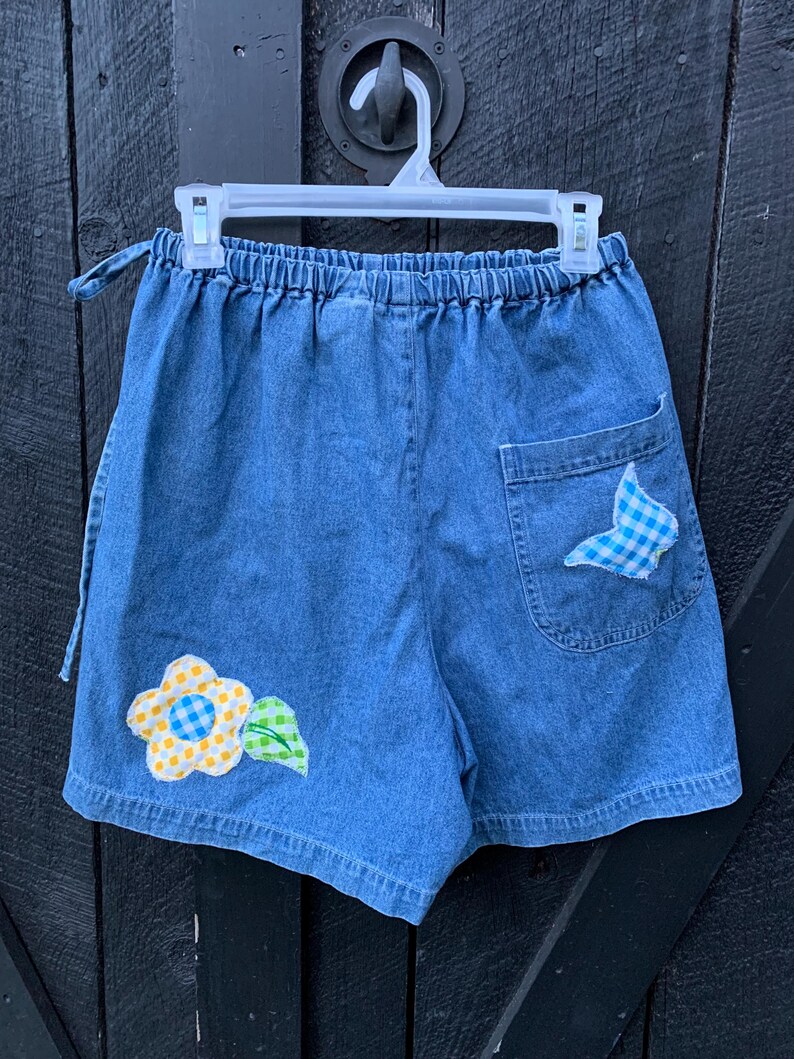 May include: Blue denim shorts with a yellow flower and green leaf applique on the left leg and a blue and white checkered fabric applique on the right back pocket.