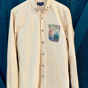 Puede incluir: Una camisa de manga larga de color amarillo claro con un bolsillo con estampado de ciervo. La camisa tiene un cuello abotonado y una etiqueta de marca que dice "Town & Tour" con la talla "Large" impresa debajo.