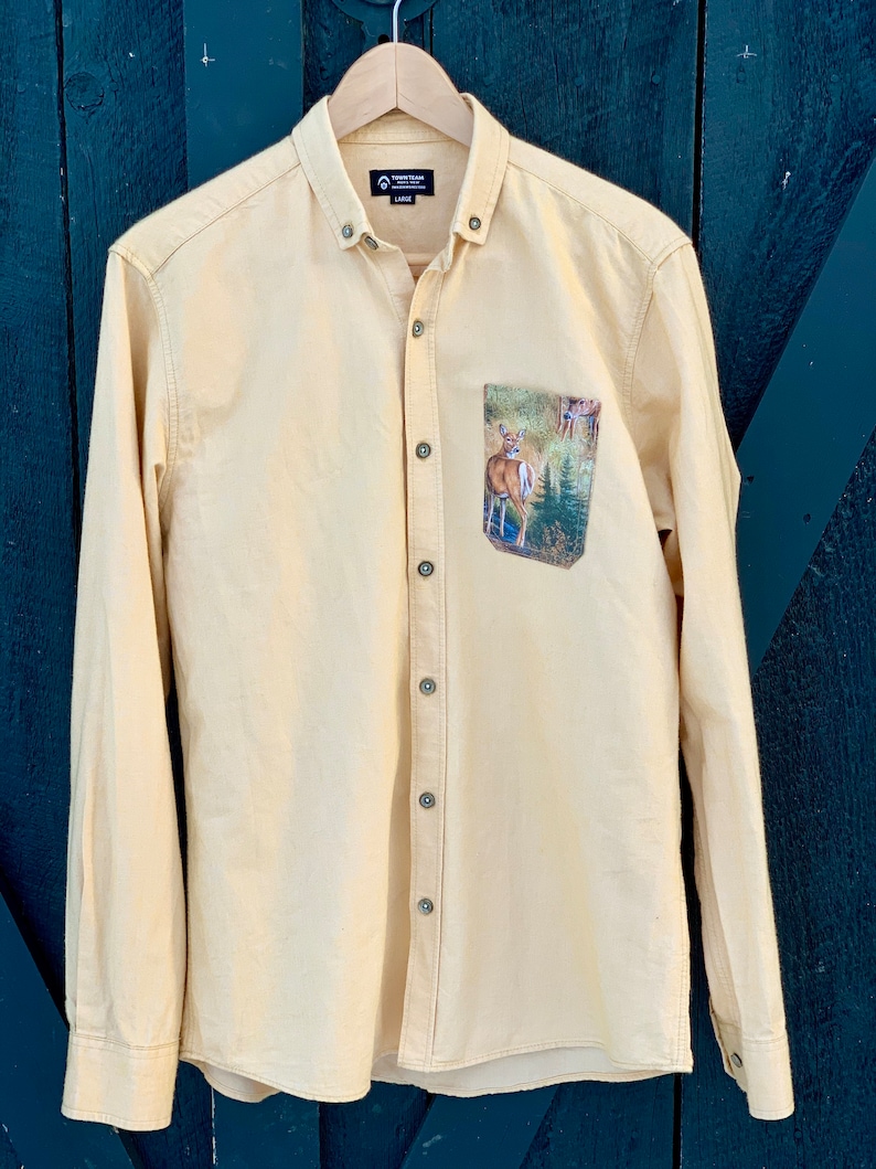 Puede incluir: Una camisa de manga larga de color amarillo claro con un bolsillo con estampado de ciervo. La camisa tiene un cuello abotonado y una etiqueta de marca que dice "Town & Tour" con la talla "Large" impresa debajo.
