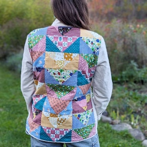 May include: A person wearing a white shirt with a patchwork quilt vest. The vest is made of various colorful fabrics with floral and geometric patterns. The person is wearing blue jeans and standing in a grassy area.