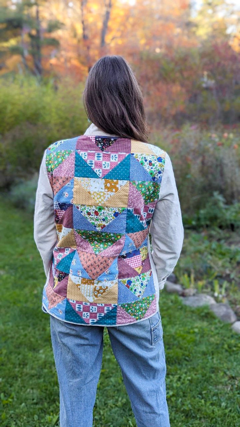 May include: A person wearing a white shirt with a patchwork quilt vest. The vest is made of various colorful fabrics with floral and geometric patterns. The person is wearing blue jeans and standing in a grassy area.