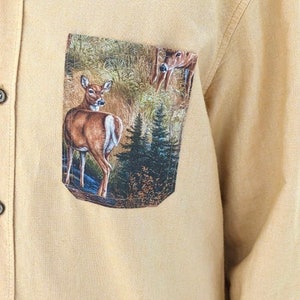 Puede incluir: Una camisa de botones de color beige con un bolsillo de parche que presenta un dise&ntilde;o impreso de dos ciervos en un entorno de bosque.