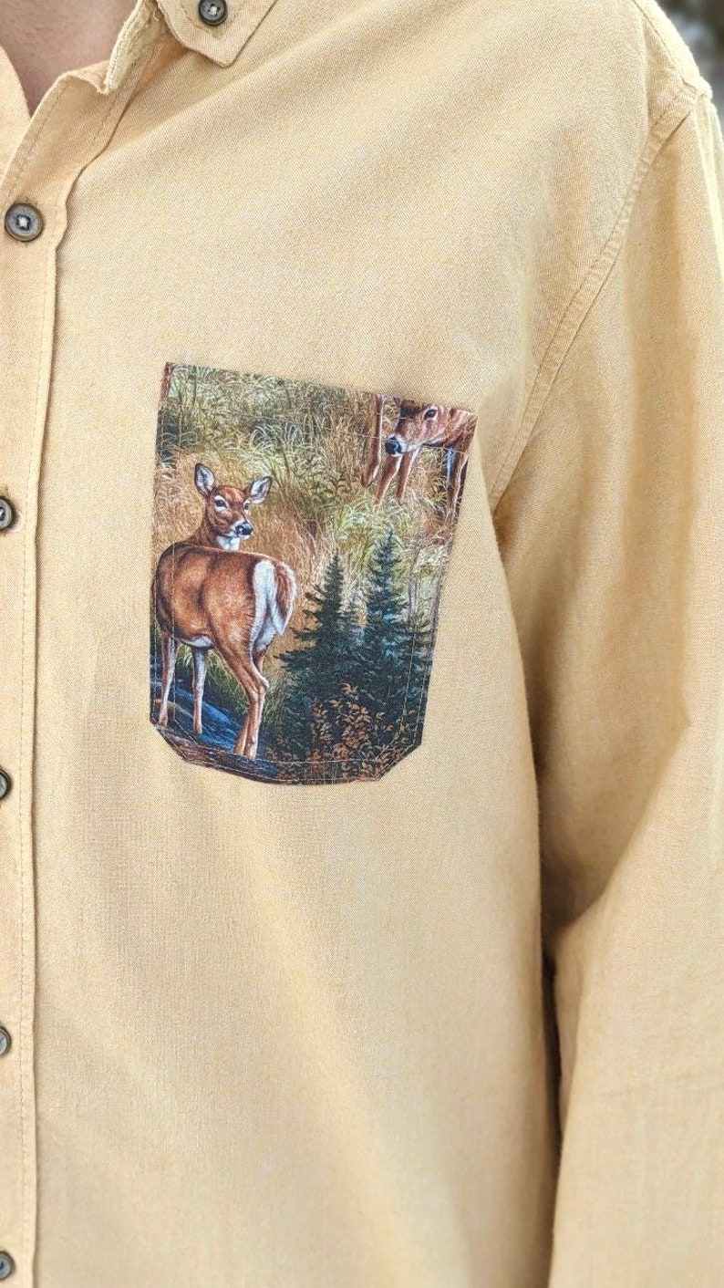 Puede incluir: Una camisa de botones de color beige con un bolsillo de parche que presenta un dise&ntilde;o impreso de dos ciervos en un entorno de bosque.