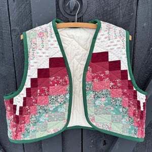 May include: A patchwork quilted vest featuring a mix of red, pink, and green floral fabrics. The vest has a cream-colored lining and is edged with green binding. It is displayed on a wooden hanger against a dark backdrop.