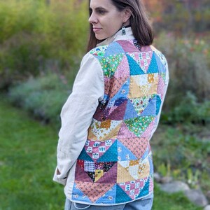 May include: A woman wearing a cream colored jacket with a patchwork quilt design on the back. The quilt is made of various colorful fabrics with floral and geometric patterns. She is wearing blue jeans.