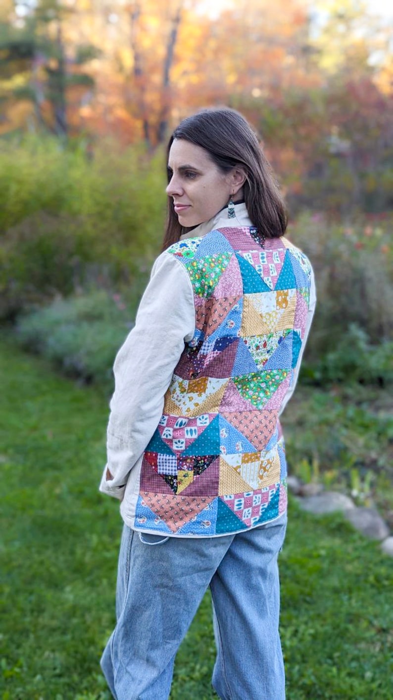 May include: A woman wearing a cream colored jacket with a patchwork quilt design on the back. The quilt is made of various colorful fabrics with floral and geometric patterns. She is wearing blue jeans.