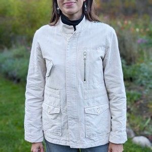 May include: A woman wearing a light beige jacket with a zipper and pockets, blue jeans, and earrings. She is standing in front of a blurred background of trees and foliage.