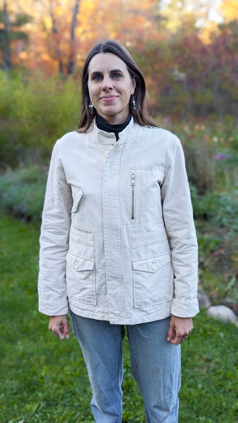 May include: A woman wearing a light beige jacket with a zipper and pockets, blue jeans, and earrings. She is standing in front of a blurred background of trees and foliage.