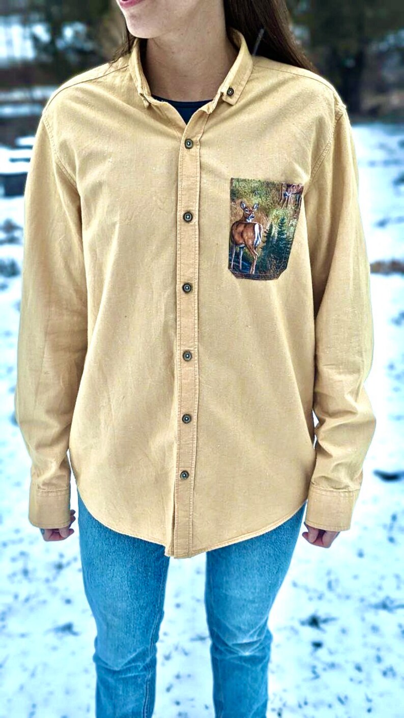 Puede incluir: Una camisa de manga larga de color beige claro con botones y un bolsillo con estampado de ciervo. La camisa tiene un corte holgado y est&aacute; hecha de una tela suave y c&oacute;moda.