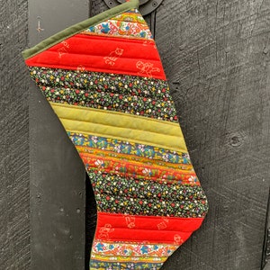 May include: A red, green, and yellow striped Christmas stocking with a quilted design. The stocking is made of fabric scraps and has a variety of floral patterns. The stocking is hanging on a dark wooden door.
