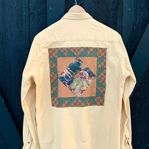 Può includere: Una camicia gialla a maniche lunghe con bottoni e un design a patchwork sul retro. Il patchwork è realizzato in tessuto marrone, verde e bianco e presenta una scena di cervi in un bosco.