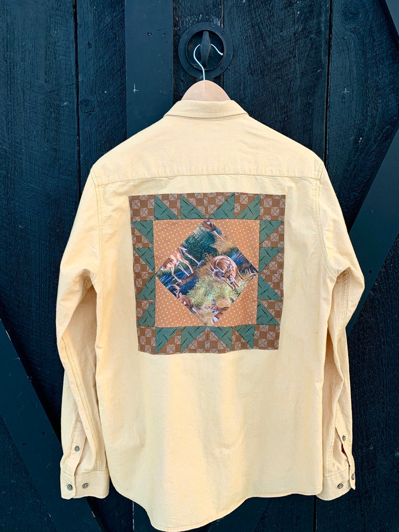 Puede incluir: Una camisa amarilla de manga larga con botones y un dise&ntilde;o de patchwork en la espalda. El patchwork est&aacute; hecho de tela marr&oacute;n, verde y blanca y presenta una escena de ciervos en un bosque.