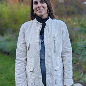 May include: A woman wearing a white jacket over a blue denim overall, standing in a grassy area with fall foliage in the background.