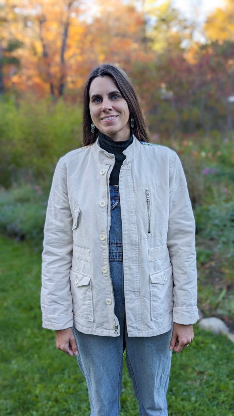 May include: A woman wearing a white jacket over a blue denim overall, standing in a grassy area with fall foliage in the background.