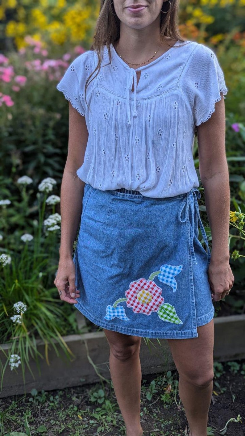 May include: A woman wearing a white eyelet top with a tie at the neck and a blue denim wrap skirt with a colorful floral applique.