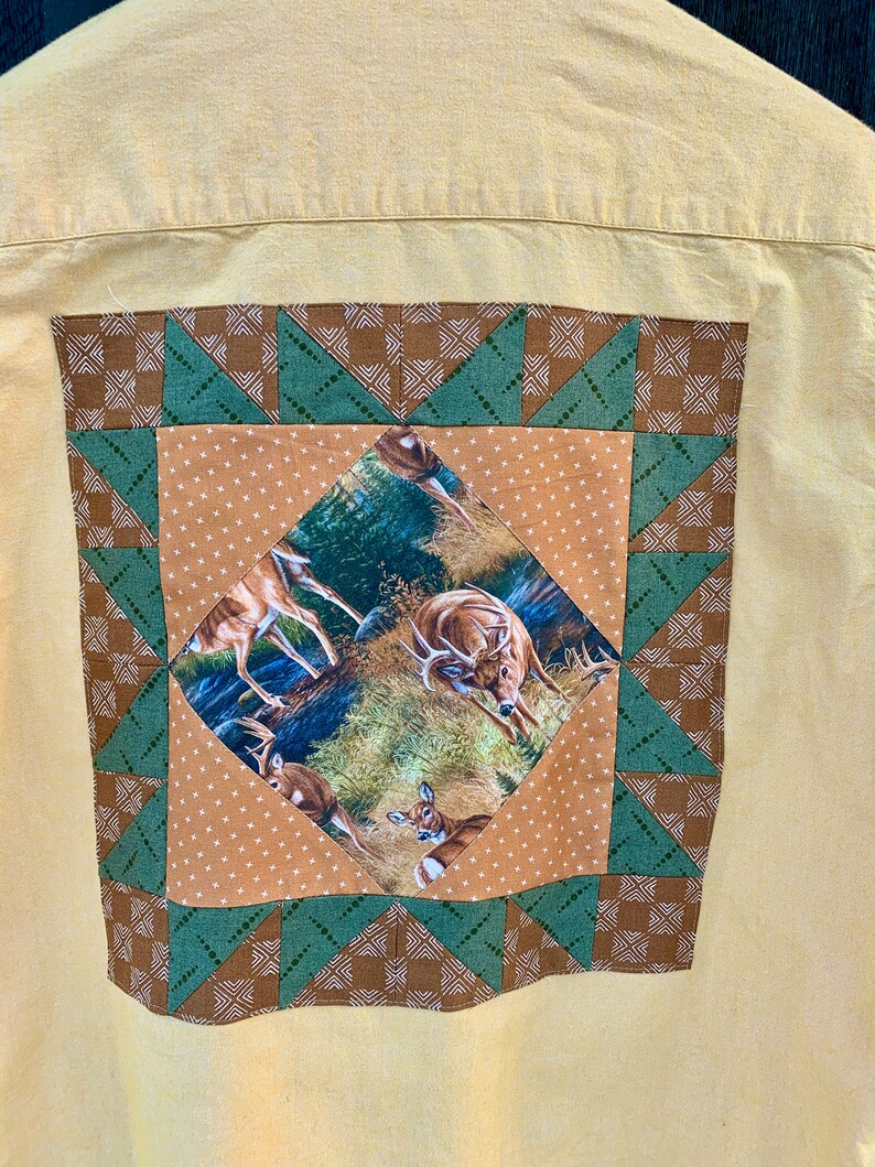 Puede incluir: Una camisa amarilla de botones con un dise&ntilde;o de patchwork marr&oacute;n y verde en la espalda. El patchwork presenta un cuadrado con un estampado de ciervo en el interior.