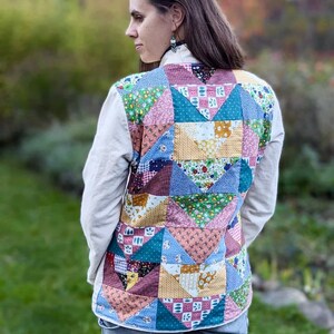 May include: A woman wearing a white shirt with a patchwork quilt vest. The vest is made of various colorful fabrics with floral patterns. The woman is wearing blue jeans.