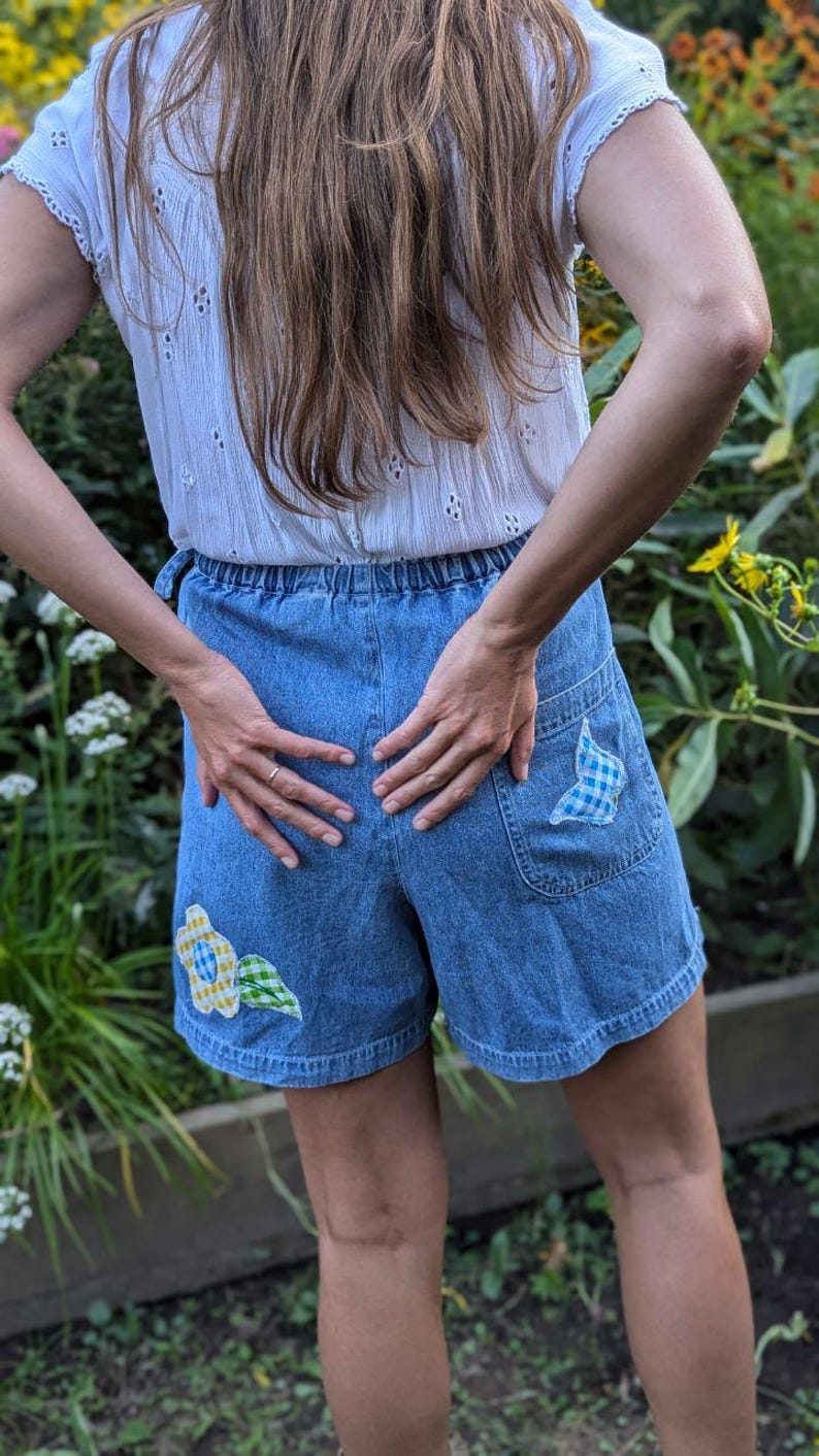 May include: A pair of blue denim shorts with a white, yellow, and blue floral patch on the left leg and a blue and white checkered patch on the right leg.