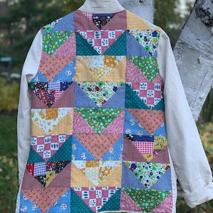 May include: A white jacket with a patchwork back made of colorful fabric squares. The squares are arranged in a geometric pattern and feature floral, geometric, and plaid designs.