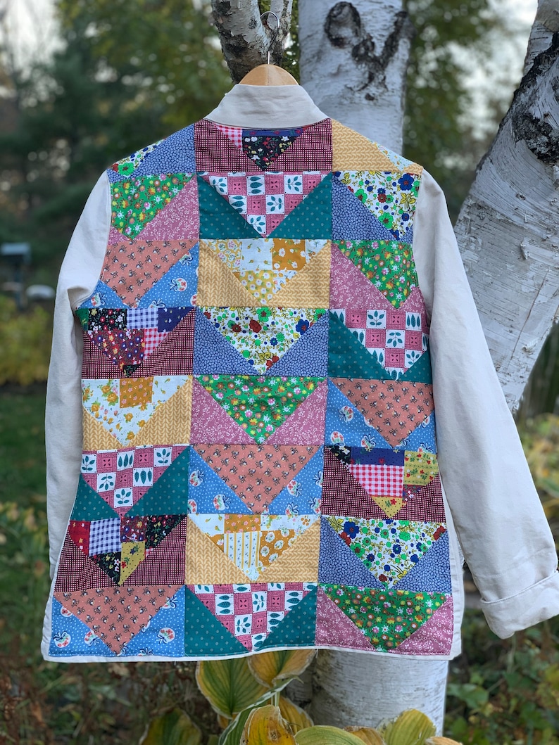 May include: A white jacket with a patchwork back made of colorful fabric squares. The squares are arranged in a geometric pattern and feature floral, geometric, and plaid designs.