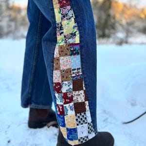 Peut inclure: Jean bleu avec un motif patchwork sur le côté. Le patchwork présente divers motifs floraux et géométriques dans les tons de rouge, jaune, bleu et marron. Des bottes marron sont visibles en bas.
