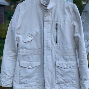 May include: A white cotton jacket with a zipper closure and multiple pockets. The jacket has embroidered details on the shoulders and the Ralph Lauren Polo Jeans Company logo on the left chest.