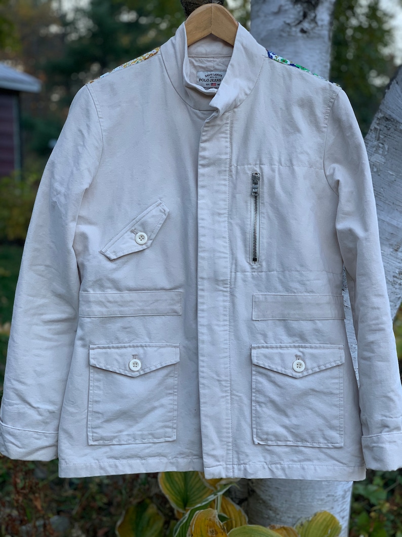 May include: A white cotton jacket with a zipper closure and multiple pockets. The jacket has embroidered details on the shoulders and the Ralph Lauren Polo Jeans Company logo on the left chest.