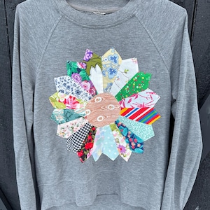 May include: A grey sweatshirt featuring a patchwork flower design. The flower is made from various fabric scraps in floral, striped, and patterned designs. The sweatshirt has long sleeves and a crew neck.