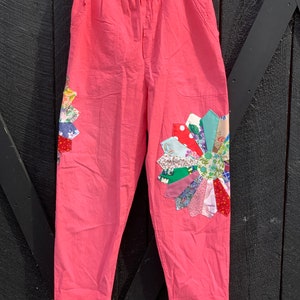 Peut inclure: Pantalon rose avec un motif de fleur en patchwork sur la jambe droite. La fleur est faite de différents tissus colorés.