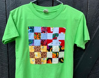 May include: A lime green t-shirt with a colorful patchwork square design on the front. The patchwork is made of different fabrics with various patterns, including floral, checkered, and geometric designs.
