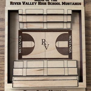 May include: A wooden sign with the text "JERRY SCHAFFER GYMNASIUM" and "HOME OF THE RIVER VALLEY HIGH SCHOOL MUSTANGS". The sign also includes a basketball court with the text "MUSTANGS" on each side and the letters "R" and "V" in the center. The sign also includes the text "THREE OAKS, MICHIGAN".