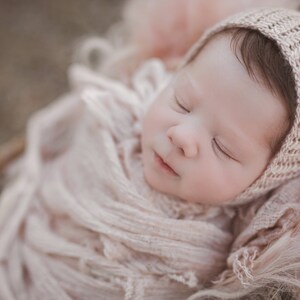 May include: A sleeping baby wrapped in a soft, light pink blanket with a crocheted hat. The baby is wearing a knitted hat with a delicate pattern.