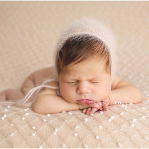 May include: A newborn baby wearing a pink knitted bonnet is sleeping on a beige knitted blanket with white dots.
