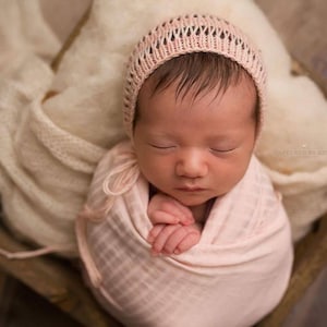 May include: A newborn baby is sleeping peacefully in a pink crocheted hat and swaddled in a pink blanket. The baby is lying in a wooden basket.