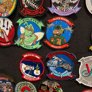 Może przedstawiać: Kolekcja haftowanych naszywek z r&oacute;żnymi odznakami jednostek wojskowych, w tym "Black Widows", "EVILEYES", "HMLA-367", "HMH-366", "VT-22 CO DET", "HMLA-3528", "VMU-4", "MALS-13", "FUELS PLATOON", "STRIKE DET", "HAMMERHEADS", "TPS 18A EXPRESS", "BYO BUDDY", "SCAREFACE" i "TEST PILOT SCHOOL".