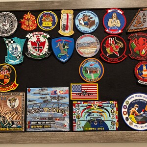 Może przedstawiać: Czarna tablica z filcu z r&oacute;żnymi kolorowymi naszywkami. Naszywki przedstawiają wojskowe odznaki, samoloty i zabawne powiedzenia. Niekt&oacute;re z naszywek to "Giant Slam Clickers", "We Slammed It So Hard", "Back Porch 1,000 Hours", "COVID Combat Cruise", "What You Do Matters Westpac 2022", "CVN Rimpac 2022" i "COVID Cruise 2020 You Got Nimitz Two Teams One Fight".