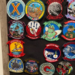 Może przedstawiać: Kolekcja kolorowych haftowanych naszywek z r&oacute;żnymi motywami wojskowymi i lotniczymi. Naszywki są umieszczone na czarnym tle w drewnianej ramie. Niekt&oacute;re z naszywek zawierają tekst, taki jak "National Test Pilot School", "Scarface", "Evileyes", "Black Wit", "VT-21 CO Dat", "Key West 21-10", "Fightin' Rhino Viper", "It's Really Quite Simple" i "HMLA-3528".