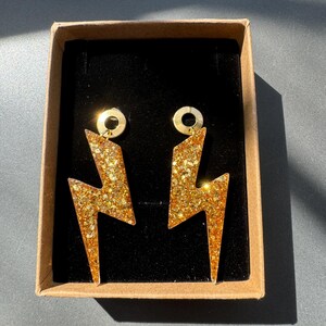 Peut inclure: Une paire de boucles d'oreilles en forme d'éclair dorées à paillettes, présentées dans une boîte en carton marron avec un intérieur noir. Les boucles ont un anneau doré pour l'attache.