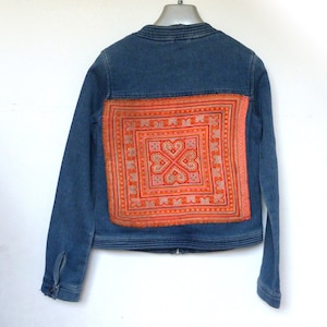 May include: A blue denim jacket with a large, colourful embroidered patch on the back. The patch features a geometric design in shades of orange, pink, and white.